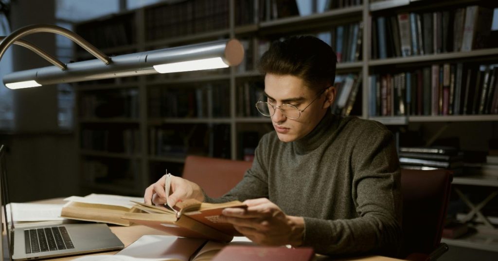 A young scholar studying in the library with books and a laptop, representing dedication and commitment to the Leo Baeck Fellowship.