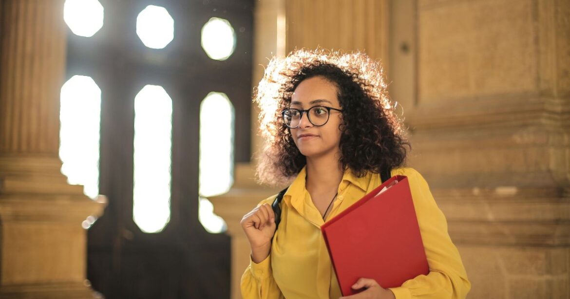GGU Worldwide Scholarship (UHUB) 2025/2026 A confident student in a yellow shirt holding a red folder, standing in a grand hallway, representing a recipient of the GGU Worldwide Scholarship (UHUB).